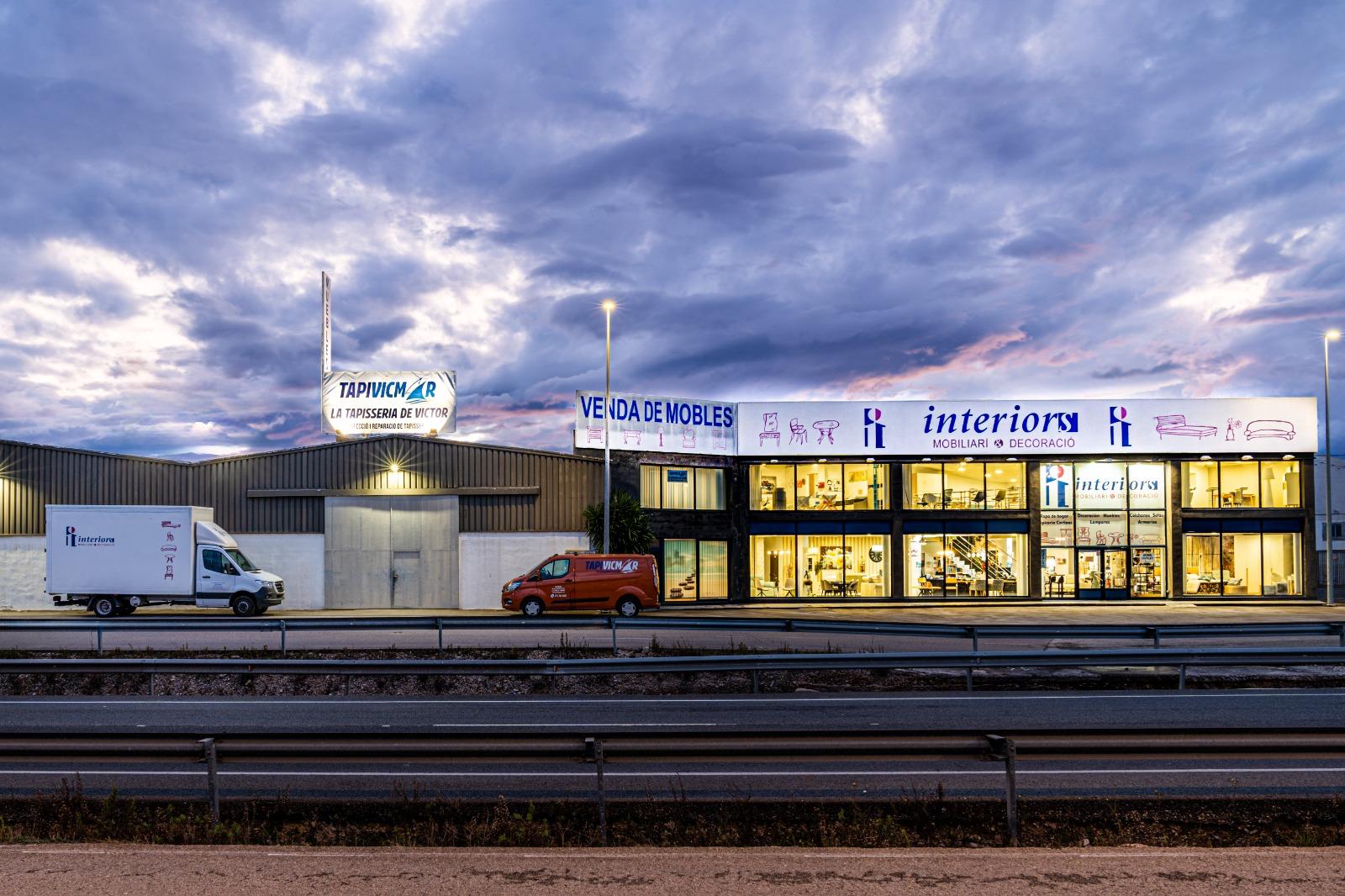 Fachada iluminada de la tienda Interiors en Vinaròs, con grandes ventanales y mobiliario de exposición.